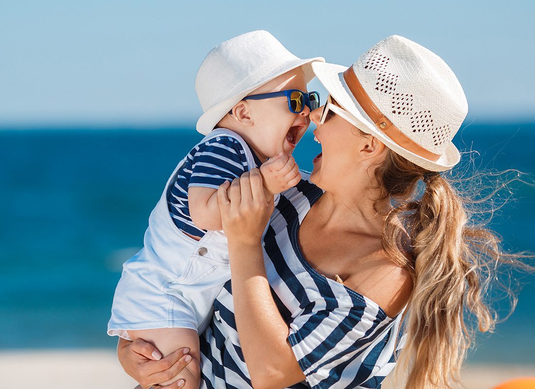 About Our Agency - Closeup View of a Cheerful Young Mother Having Fun Holding and Playing with her Baby Son on the Beach During the Summer